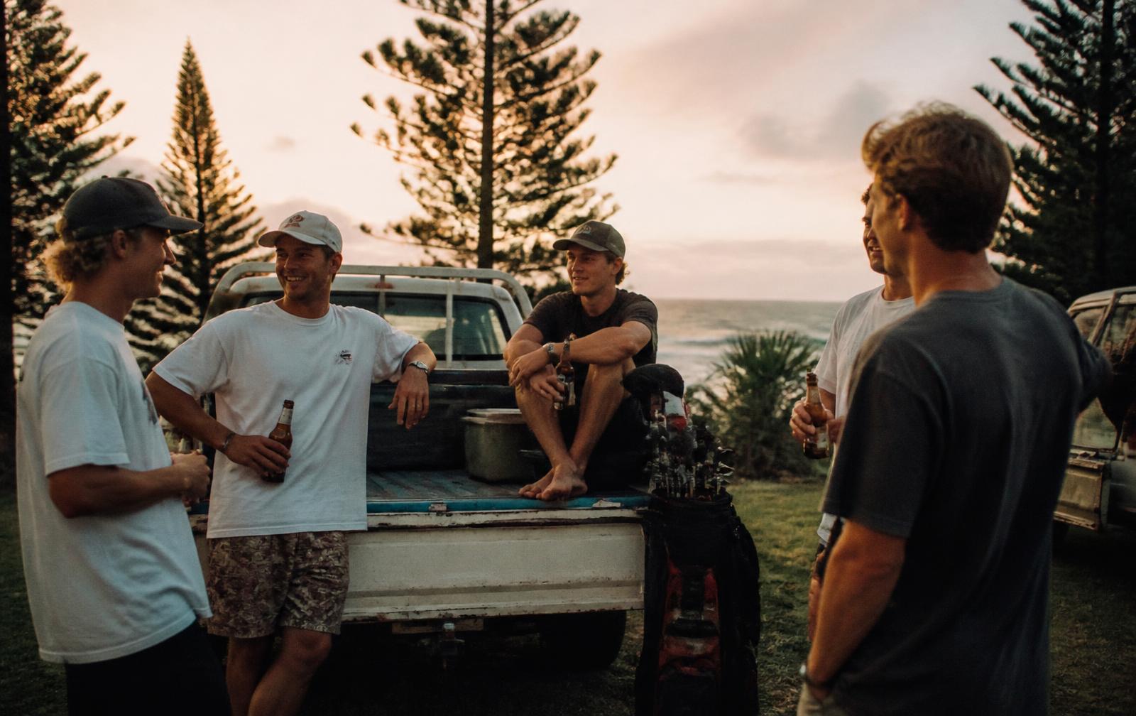 Group of young Central Coast mates at dusk around a ute tailgate with boardshorts, caps and a golf bag